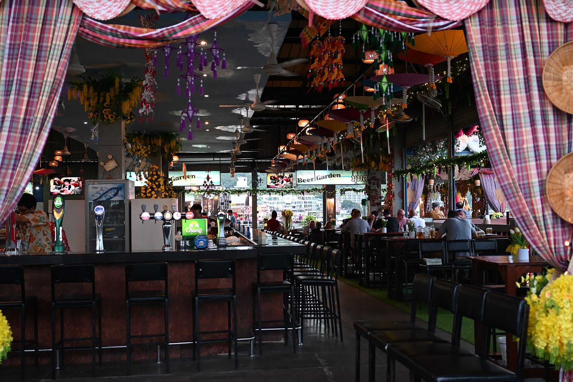 A relaxed open-air beer garden bar with high stools, hanging decorations and people enjoying evening drinks
