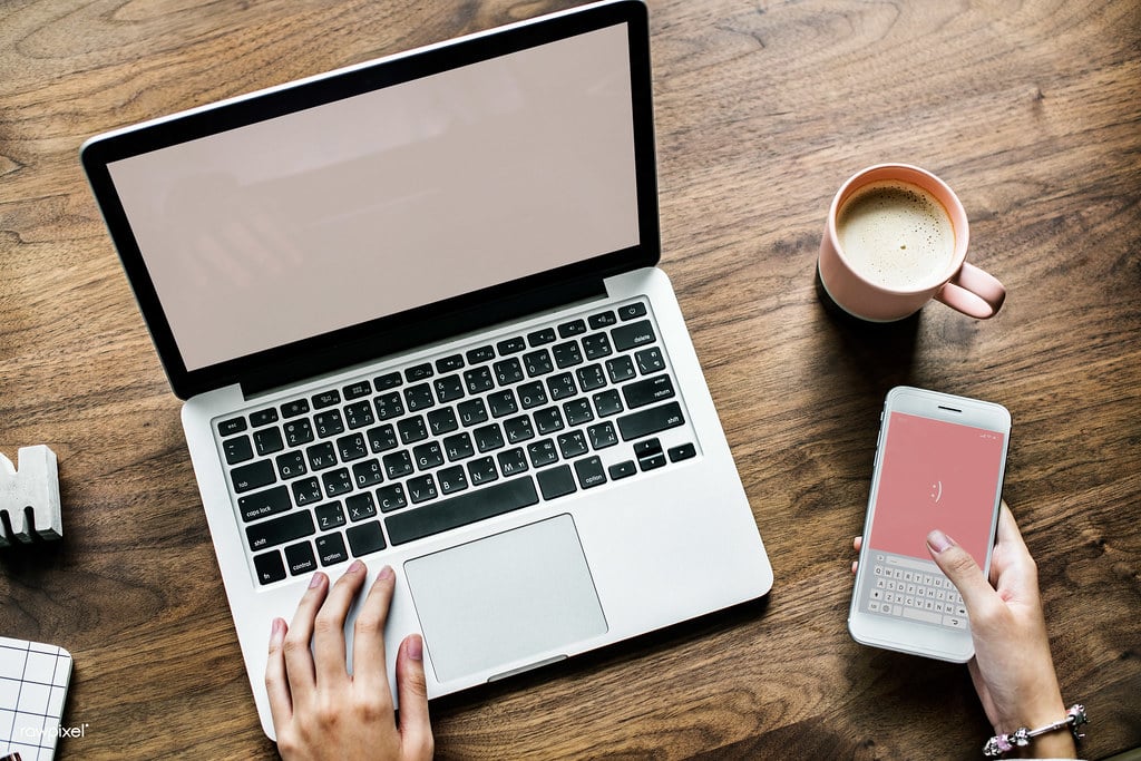 Person booking travel online at a wooden table using a laptop and smartphone with a cup of coffee