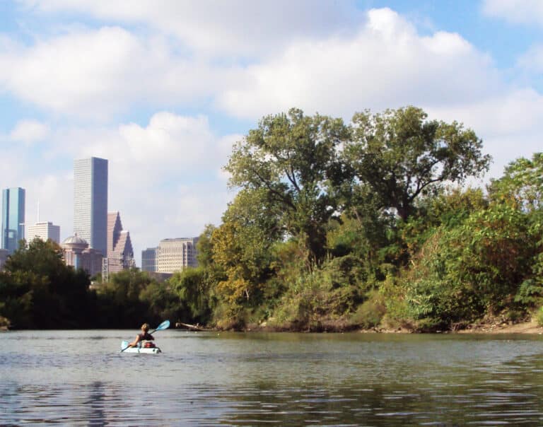 A kayaker paddling Buffalo Bayou in Houston with trees along the bank and the downtown skyline behind