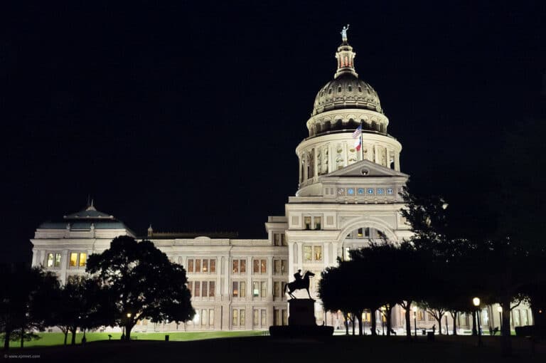 The Texas State Capitol building in Austin lit up at night, with its pink granite dome above the tree-lined grounds