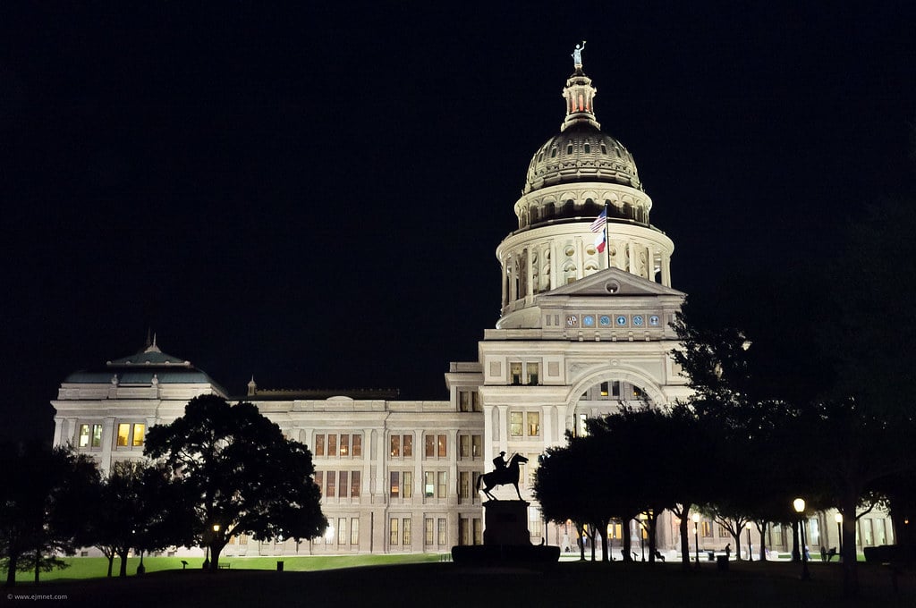 The Texas State Capitol building in Austin lit up at night, with its pink granite dome above the tree-lined grounds